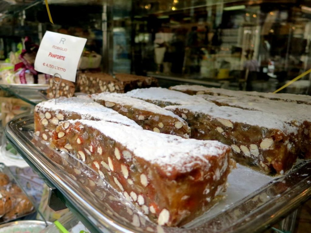 Rectangular slices of panforte dusted with powdered sugar are arranged in a glass case at an Italian bakery, price sign behind.