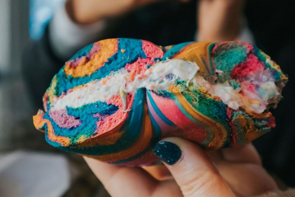 A person holding a rainbow-colored bagel with cream cheese.