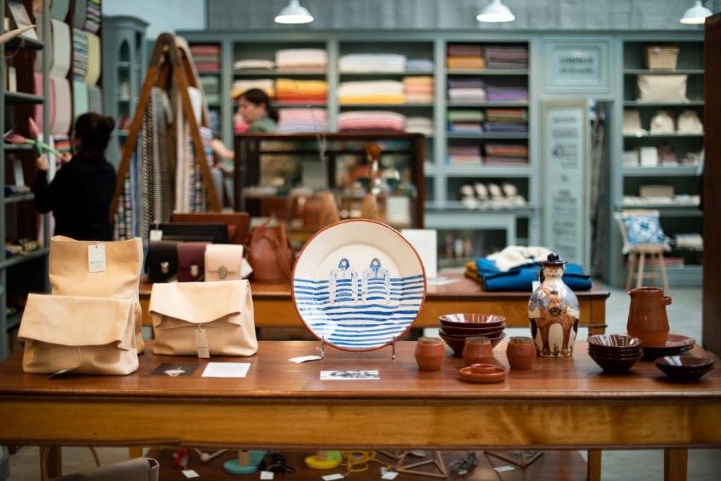 Inside a local shop in Lisbon, a display showing a blue and white plate, brown glossy ceramics and handmade bags and purses, with shoppers and shelves in the background