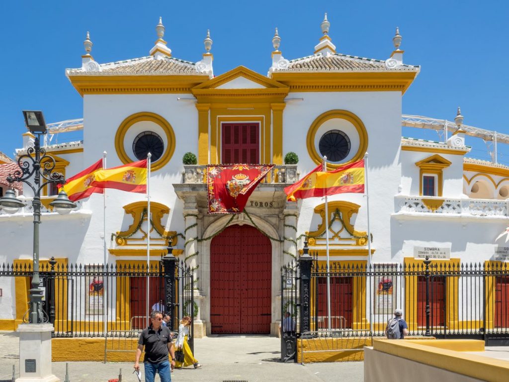 The facade of Seville’s Plaza de Toros glows under clear skies, with Spanish flags and two people walking past red doors.