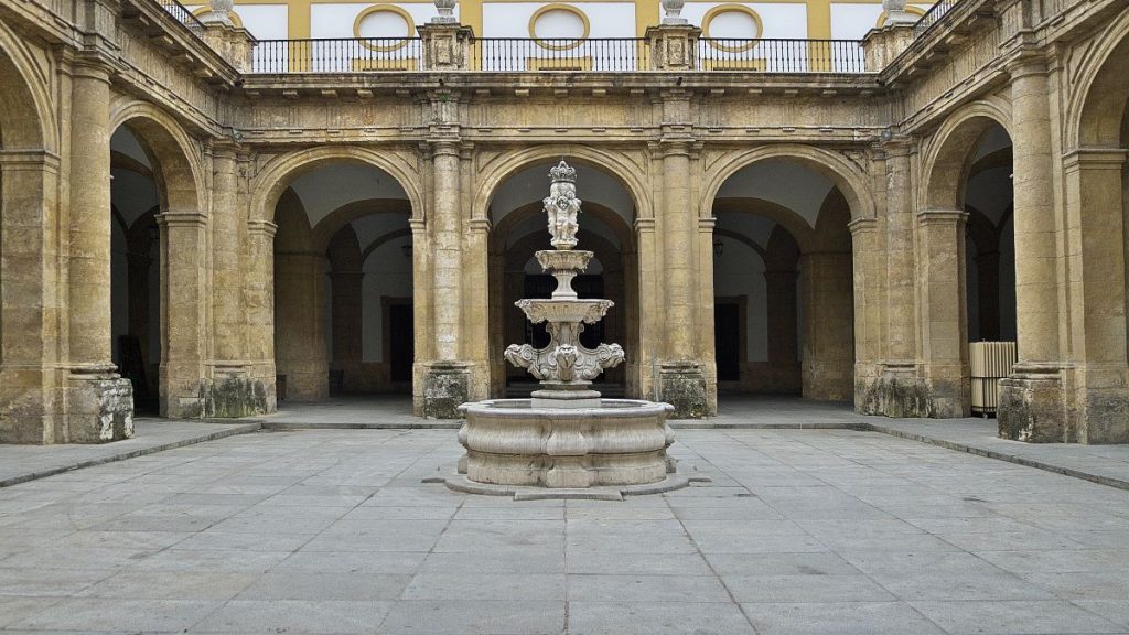 A carved stone fountain flows in a courtyard with yellow-trimmed columns and arches, evoking Sevilles historic architecture.