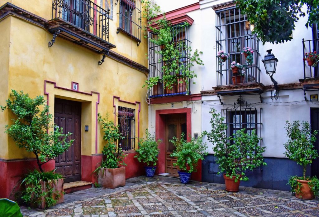 A vibrant courtyard in Seville features colorful buildings, potted plants, wrought iron balconies, and a picturesque cobblestone floor.