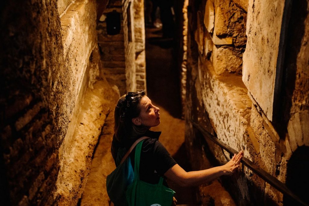 A woman with a backpack walks through a narrow, dimly lit stone corridor in ancient European architecture of the Catacombs of Rome. She is touching the stone wall.