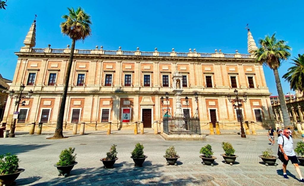 A grand historic building in Seville displays statues, tall windows, and palm trees as people walk through a sunlit plaza.