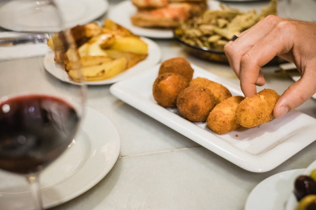 A hand reaches for a golden croquette from a white plate at a bustling Spanish Tapas bar in Barcelona, with wine and other tapas plates on the table.
