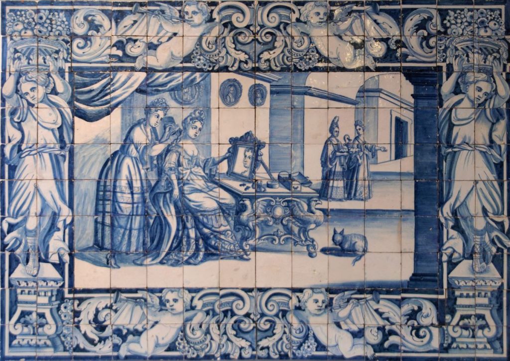 blue and white ornate tiles from Portugal, in the centre a scene of 2 ladies looking in a mirror and getting ready, within a stunning frame of cherubs and scrolls. From the National Tile museum in Lisbon