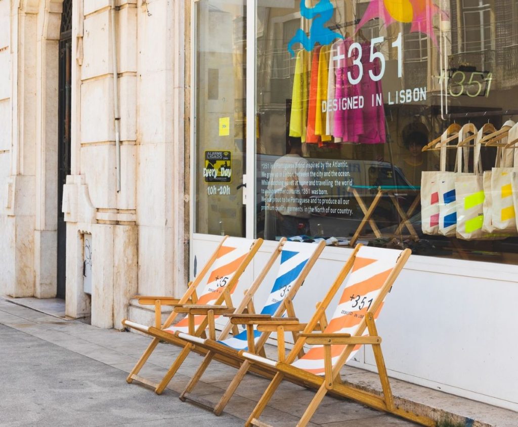 A shop front in Lisbon, with plate glass window, saying +351, designed in Lisbon, and 3 brightly coloured deckchairs outside