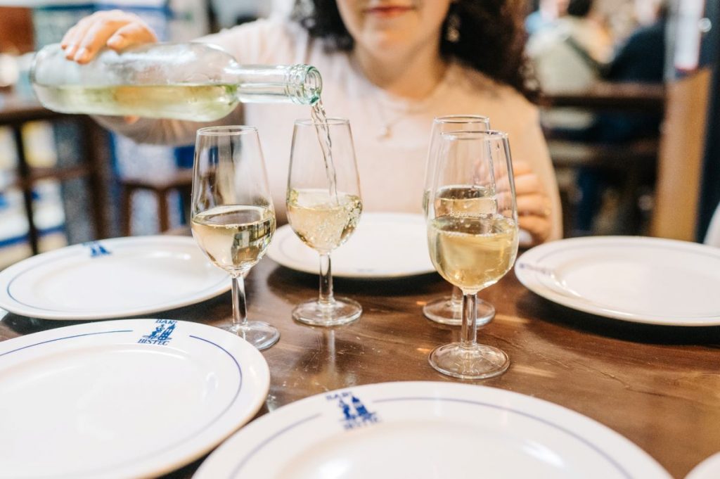 A hand pours white wine into a glass on a wooden table set for 4 people who are doing sherry tasting in Jerez, Spain.