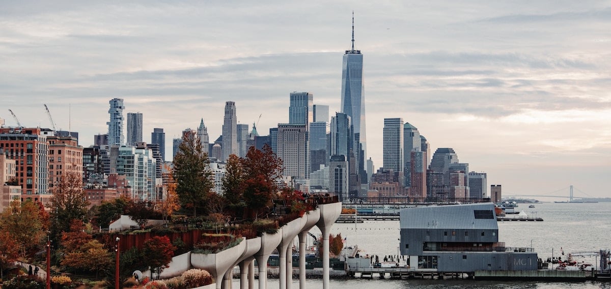 Lower Manhattan skyline with One World Trade Center centered, autumn trees on Little Island park, and the Hudson River in front.