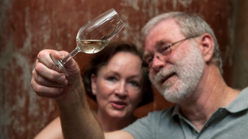 An older man with a gray beard closely examines a wine glass during a sherry tasting in Jerez, while a woman observes.