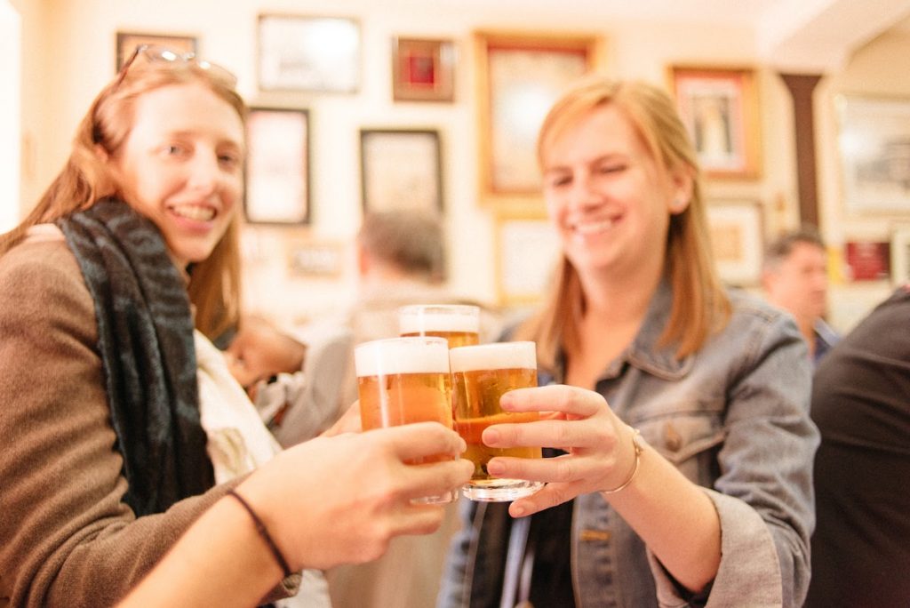 Two women smile and clink beer glasses in a bright Madrid bar, with framed pictures decorating the wall behind them.