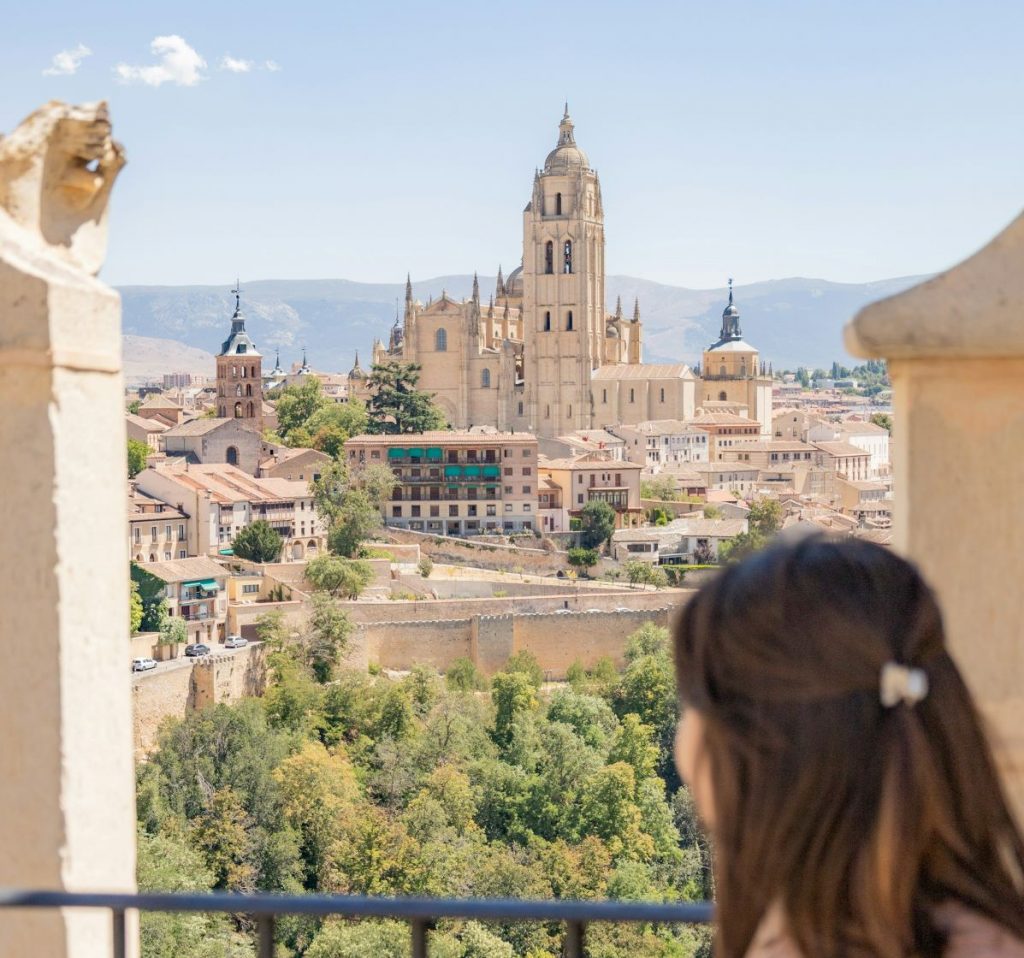 A woman with brown hair stands beneath stone archways, overlooking Segovia’s Alcazar and its Gothic cathedral under a clear sky.