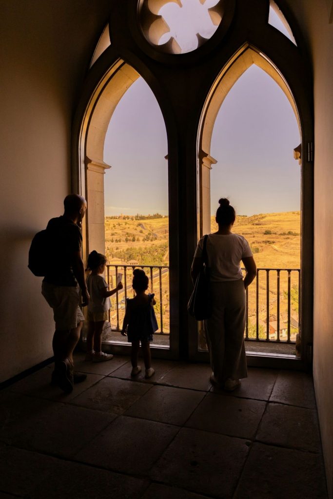 A family of four stands before ornate arched windows, gazing at sunlit hills from inside the historic Alcazar of Segovia.