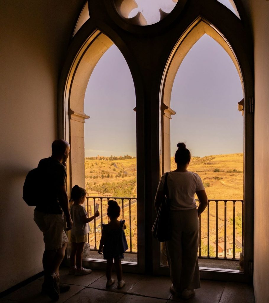 A family of four stands before ornate arched windows, gazing at sunlit hills from inside the historic Alcazar of Segovia.