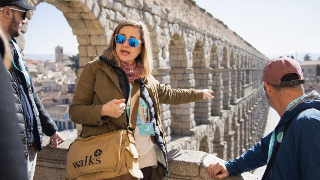 A tour guide in sunglasses gestures to a small group beside Segovias ancient aqueduct, with the Alcazar and city skyline behind.