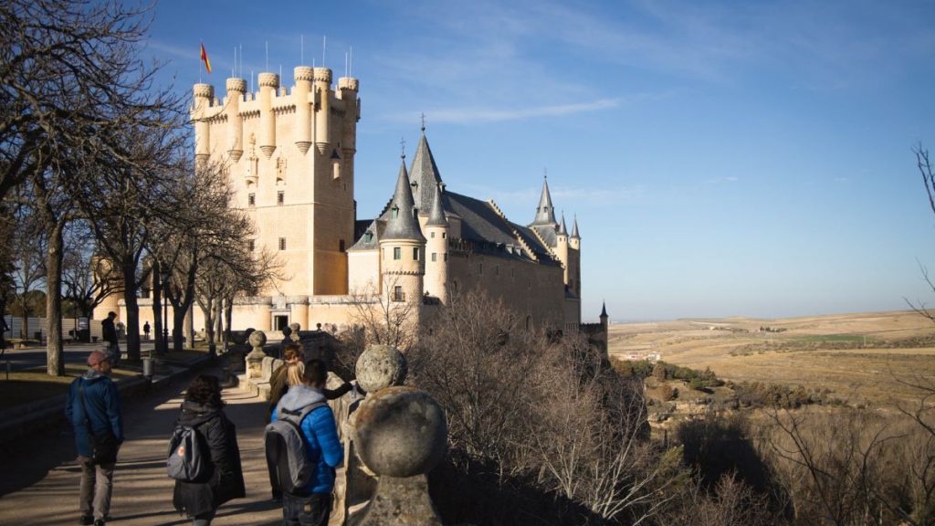 Tourists stroll on a path next to the Alcázar of Segovia, a historic Spanish castle with tall towers and scenic countryside.