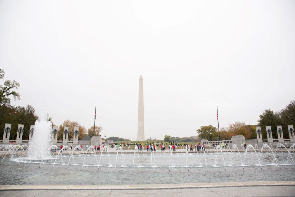 The Washington Monument rises behind a large fountain and pool, with people walking nearby under cloudy skies in Washington, D.C.
