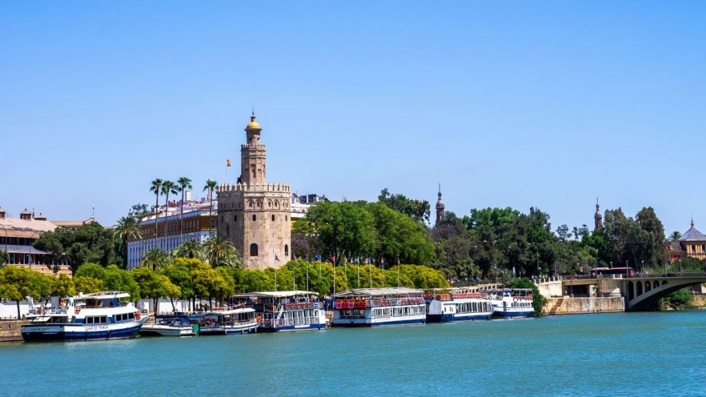 The Torre del Oro, a historic stone tower in Seville, Spain, stands beside the Guadalquivir River with boats nearby.