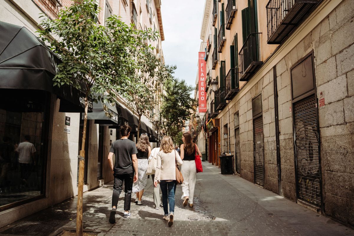 Four people stroll along a narrow, sunlit street in Madrid, passing shops, trees, and a red Flamenco sign overhead.