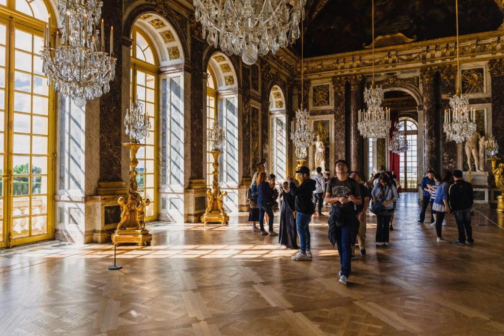 Visitors stroll through the ornate Hall of Mirrors at the Palace of Versailles, surrounded by golden decorations and shimmering chandeliers.