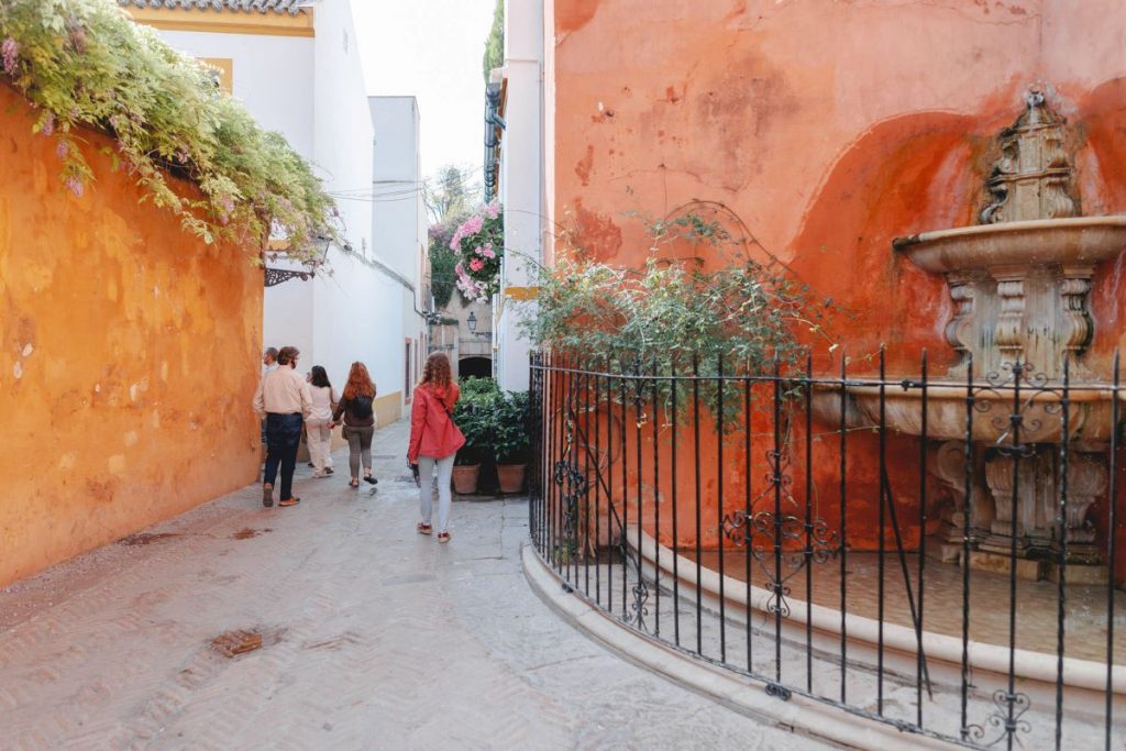 Several people stroll along a narrow street with orange walls in Seville; children play by a stone fountain beside greenery.