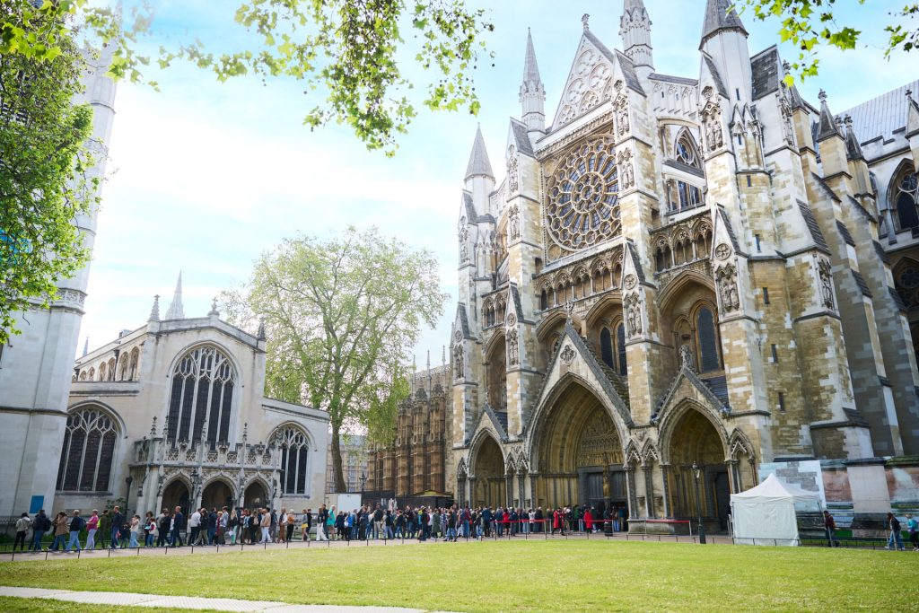 A crowd of people waits outside Westminster Abbey in London, admiring its ornate Gothic architecture and tall spires on a sunny day. One of the top locations on a London travel checklist