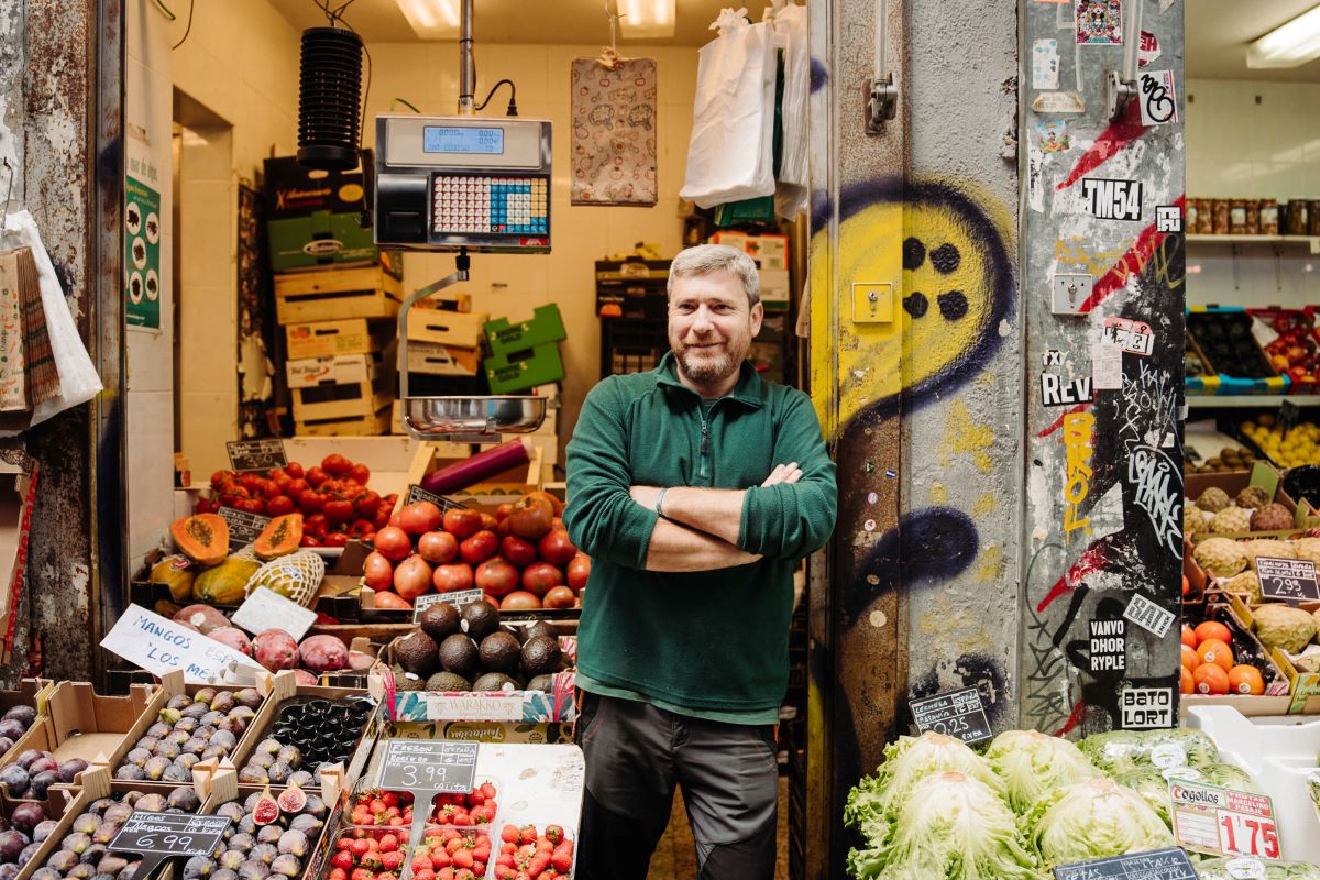 man in green shirt standing on wall in front of fruit stand in the Madrid neighborhood of Lavapies, whose name means "wash feet" 