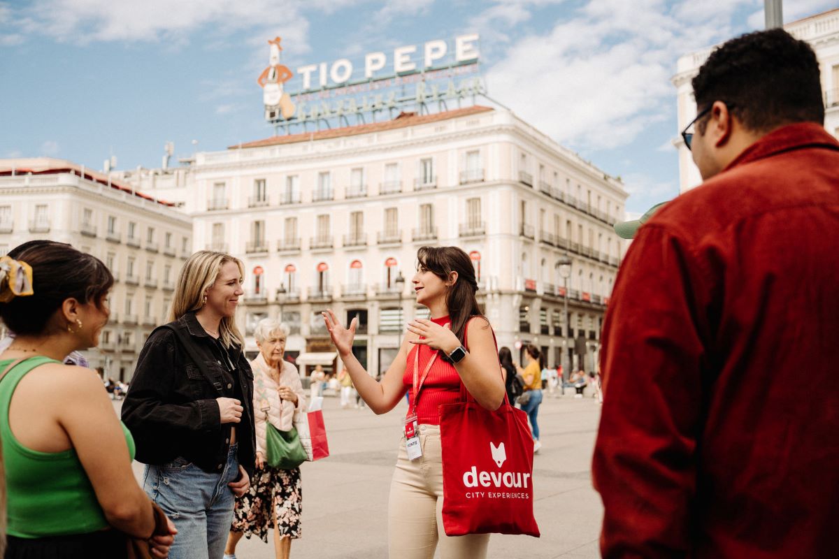 A tour guide with a red bag leads a group in Puerta del Sol, Madrid, gesturing near the Tio Pepe sign.