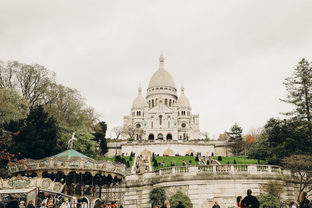 Sacré-Cœur Basilica rises on Montmartre hill in Paris, with visitors relaxing on the lawns and a vintage carousel nearby. A great place to walk to on a solo trip to Paris