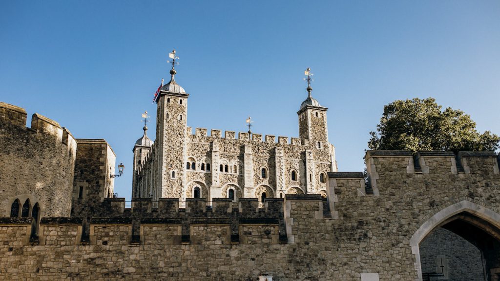 The Tower of London, located in central London, displays its stone walls and turrets beneath a clear sky beside leafy trees. one of the sights that is a must on a London travel checklist