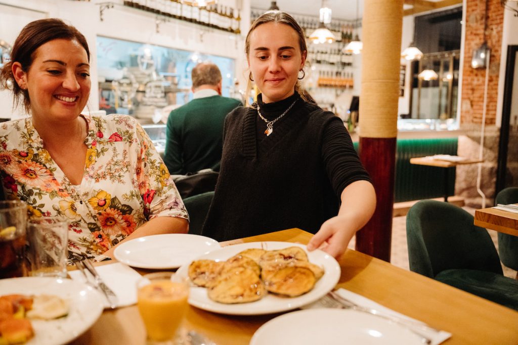 Tapas are one of the reasons why is seville worth visiting - a woman serves food to another, both enjoying the bustling and cozy dining ambiance.