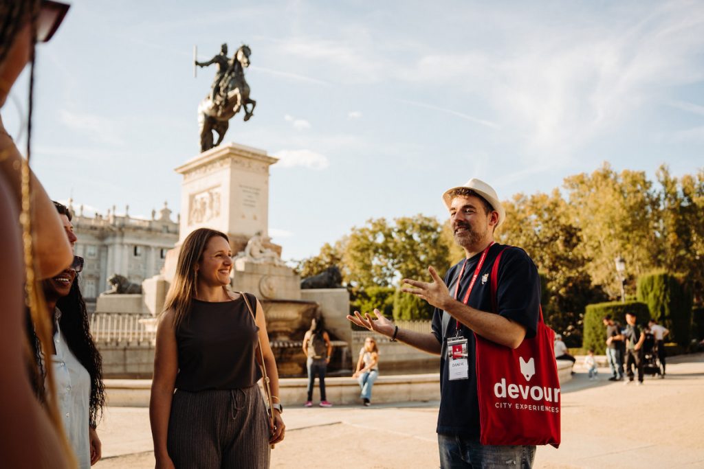 A group of tourists listen to a guide talk, with Plaza de Oriente in the background, taking a tour is one of the best ways for solo traveling madrid.
