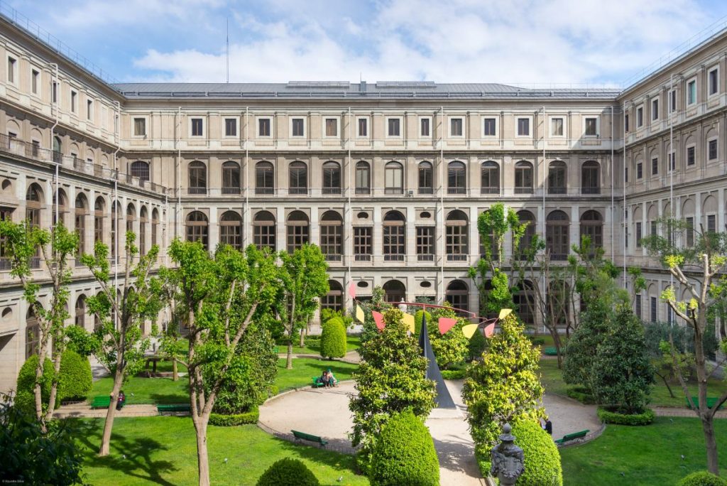 The courtyard garden at Museo Reina Sofía in Madrid features trimmed trees, benches, walking paths, and colorful red-and-yellow banners.