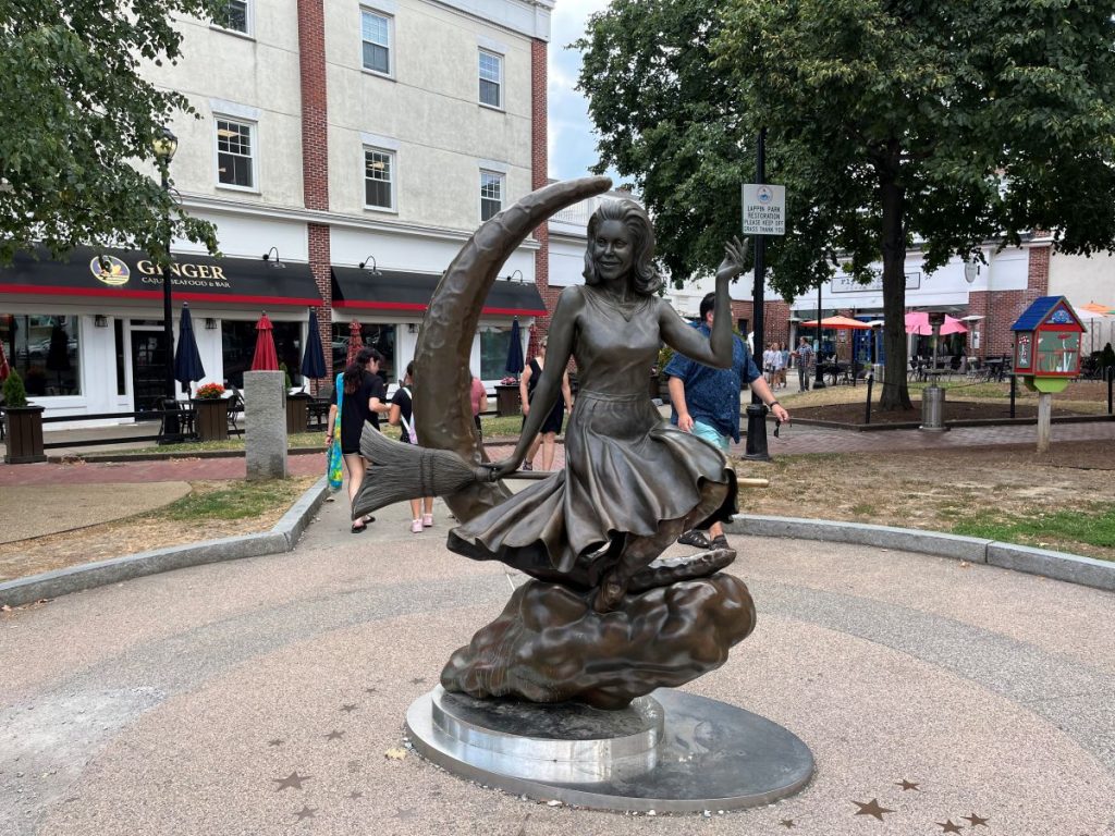 A bronze statue of a woman riding a crescent moon stands in Lappin Park, Salem, as visitors walk and relax nearby.