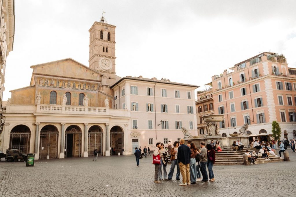 Several people gather by a fountain in Romes Piazza Navona, with historic buildings and a clock tower nearby.