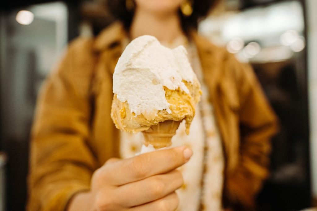 A person in a brown jacket joyfully holds a vanilla and caramel ice cream cone, with a blurred indoor setting.