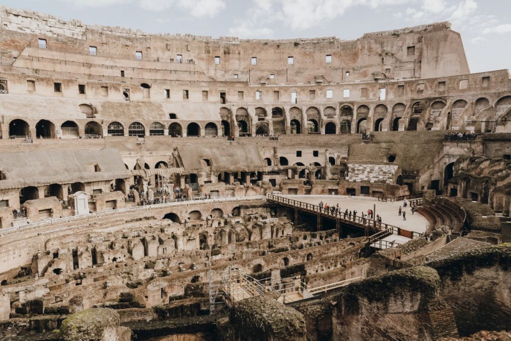 Visitors explore the Roman Colosseum in Rome, strolling a wooden path amid tiered seating under a partly cloudy sky.