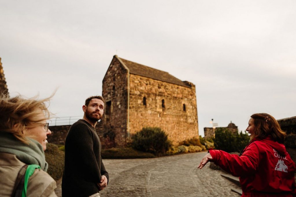 Three people converse on the Royal Mile in Edinburgh; a woman in a red jacket gestures energetically while others listen attentively.