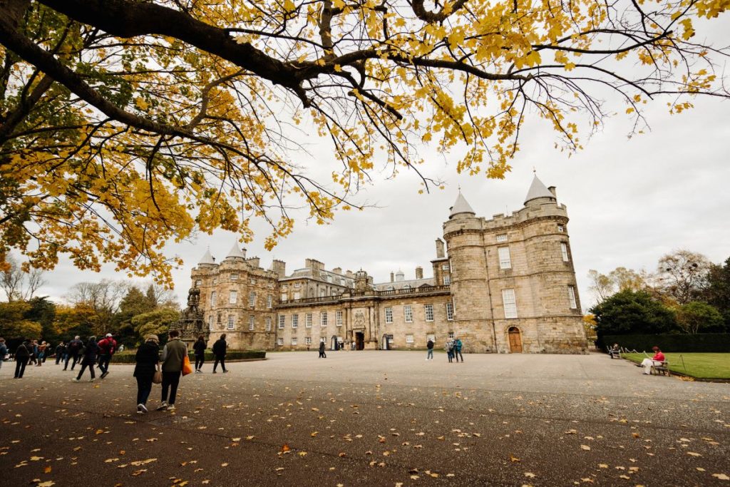 Edinburgh Castle stands atop Castle Rock, surrounded by yellow-leaved trees and people walking along the esplanade on an overcast day.