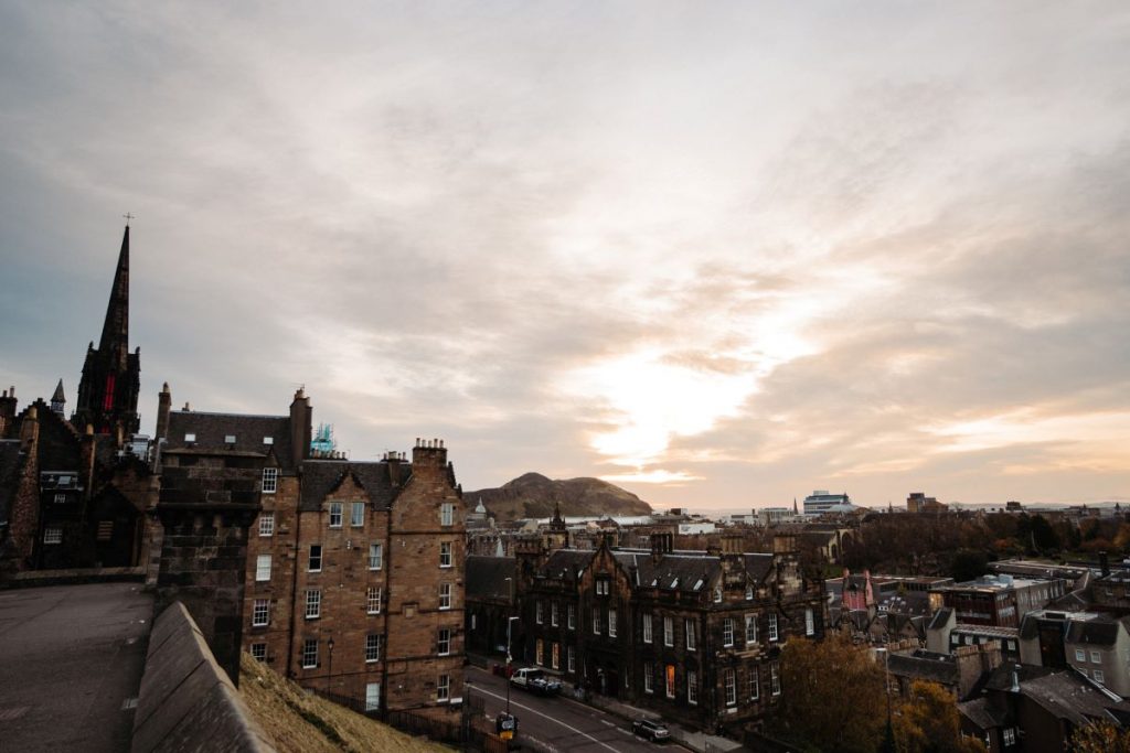 Tall stone buildings and the steeple of St. Giles Cathedral line Victoria Street in Edinburgh at sunset, with Arthur’s Seat behind.