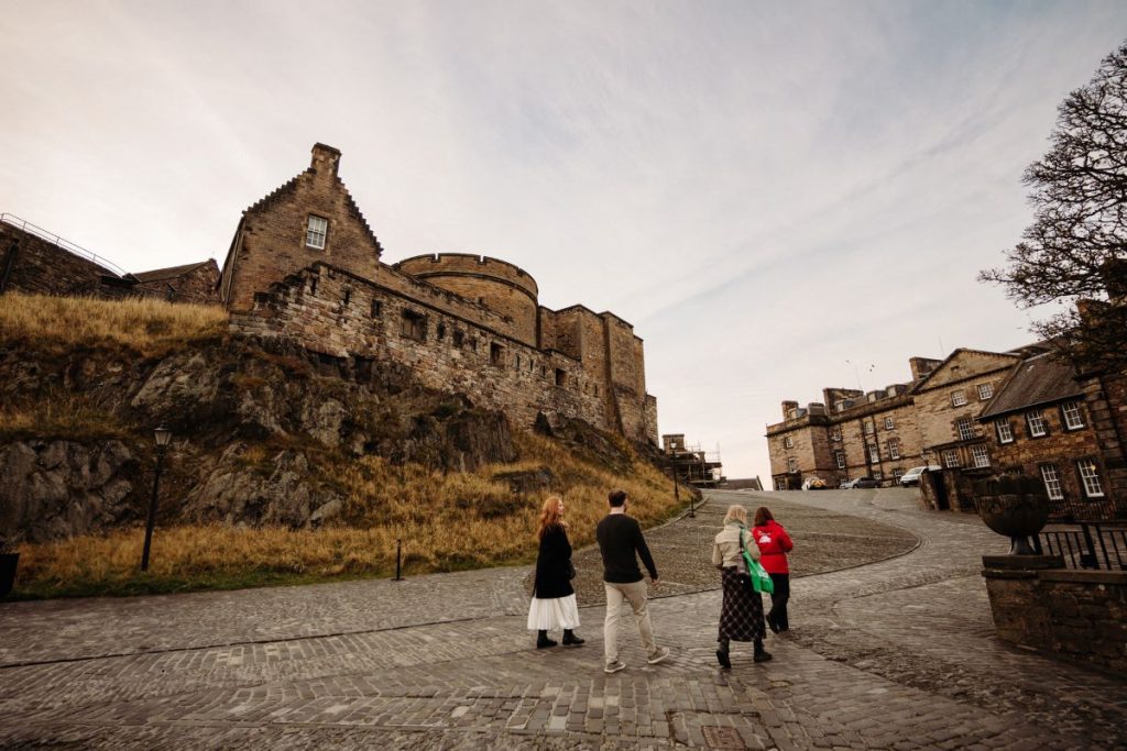 Four people walk up the cobblestone path toward Edinburgh Castle, surrounded by grassy hills and historic stone buildings under cloudy skies.