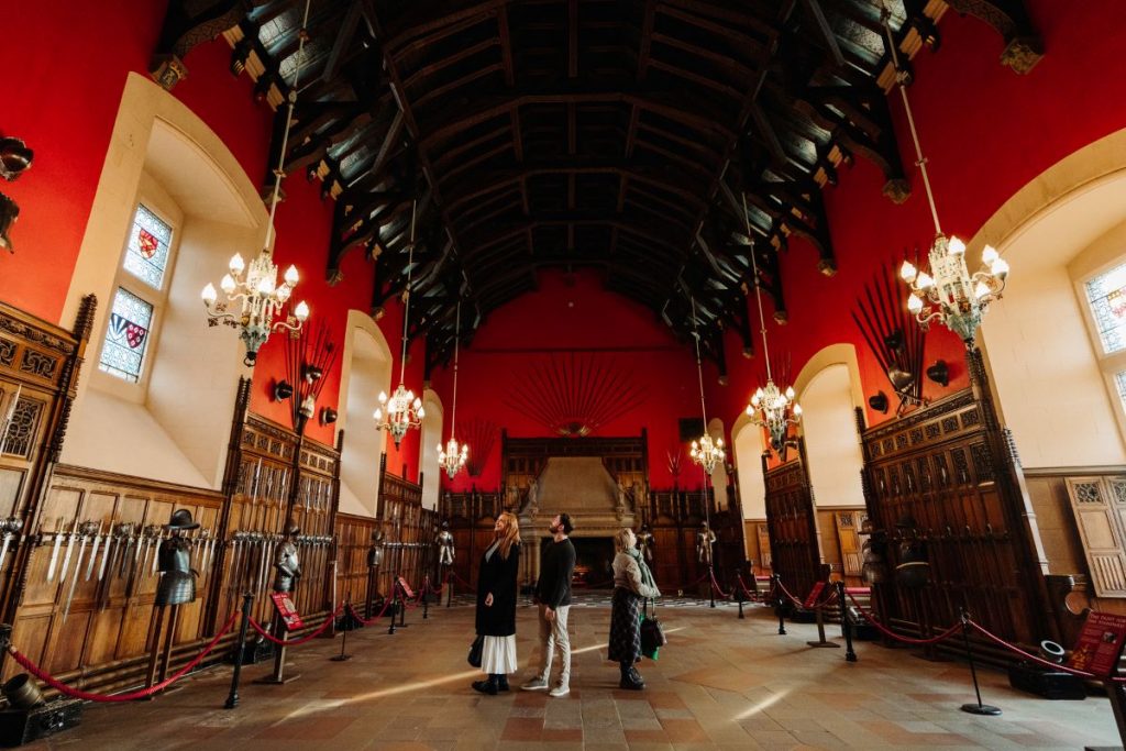 Three people stand in the Great Hall of Edinburgh Castle, surrounded by red walls, stained glass windows, and medieval armor displays.