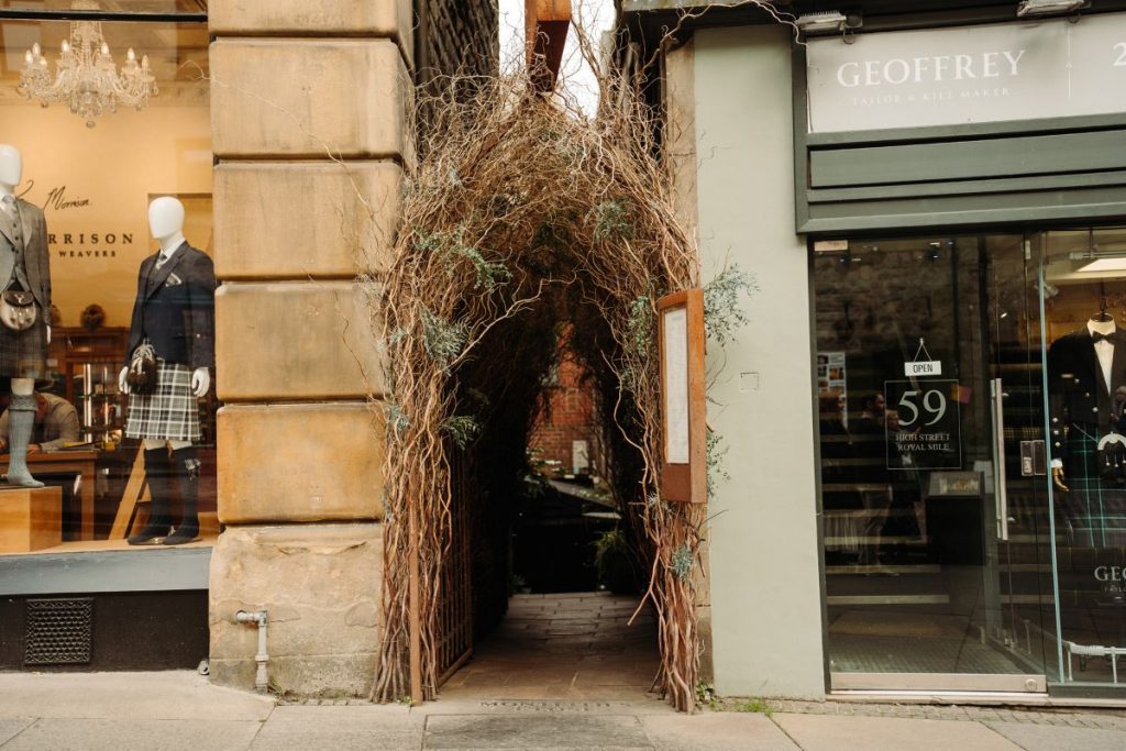 A rustic archway of branches leads to Advocates Close off Edinburgh’s Royal Mile, inviting exploration between traditional clothing shops.