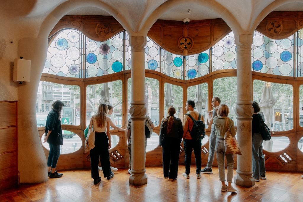 Several people stand inside Casa Batlló, Barcelona, admiring the city view through Gaudí’s colorful stained glass and curved wooden windows.