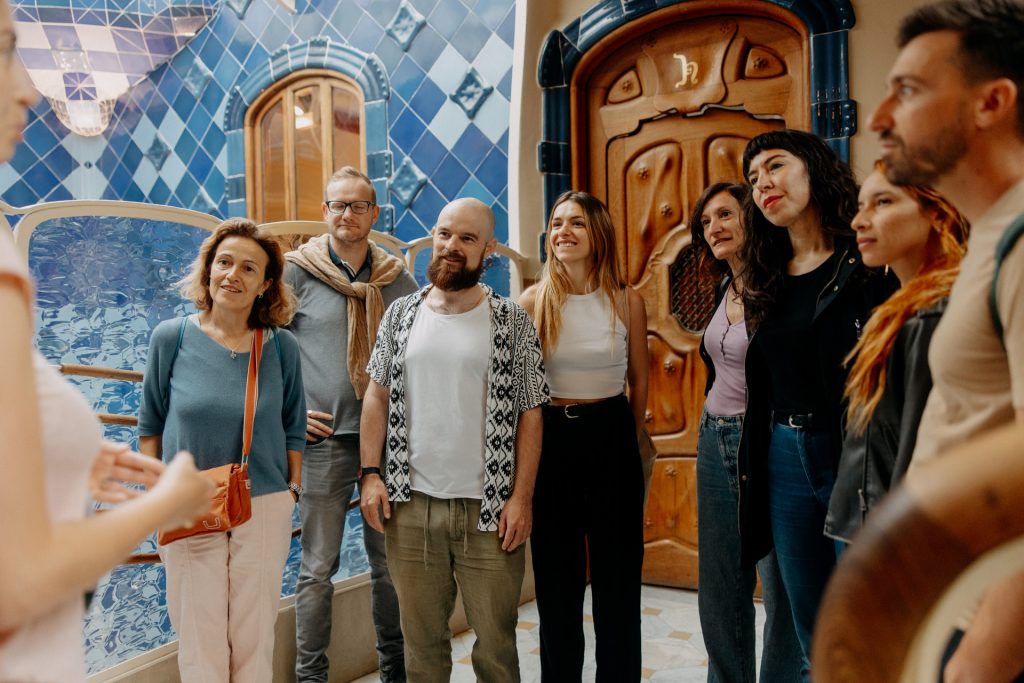 A cheerful group listens to a guide inside casa Batllo, surrounded by blue azulejos and an ornate wooden door.