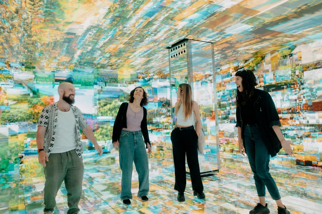 Four people smile and laugh together in a brightly lit room, surrounded by colorful, floor-to-ceiling digital projections, one of the experiences inside casa Batllo in Barcelona
