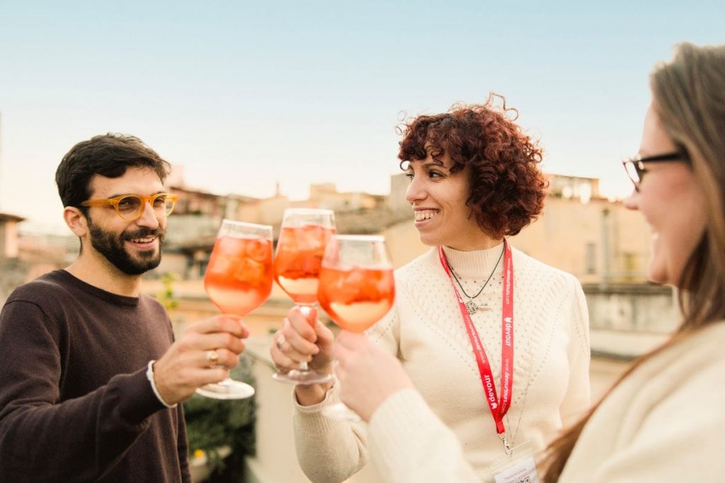 Three people toast with orange Aperol spritz drinks, smiling in front of Milan city buildings during a self-guided tour.