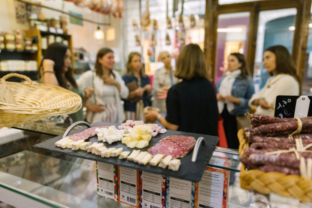 Several people chat beside a deli counter with assorted cheeses and cured meats; shelves and hanging meats create a lively Parisian atmosphere. Enjoy a host of french foods on your solo trip to Paris.
