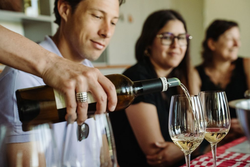 A person serves white wine into glasses at a small dining table as three friends smile and enjoy conversation together.