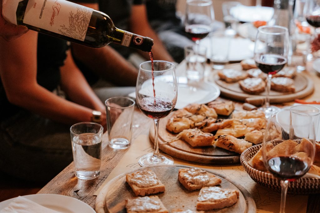 A tbale showing the key food and wine to try for a taste of Tuscany. A hand pours red wine into a glass on a Tuscan table surrounded by breads, pastries, and friends enjoying a meal.